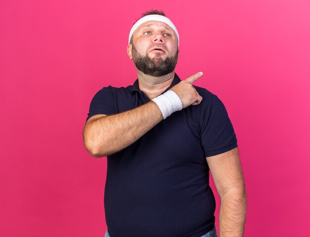 sad adult slavic sporty man wearing headband and wristbands pointing at side isolated on pink wall with copy space