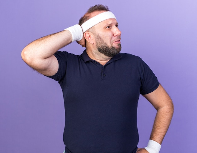 Free photo sad adult slavic sporty man wearing headband and wristbands putting hand on head behind and looking at side isolated on purple wall with copy space