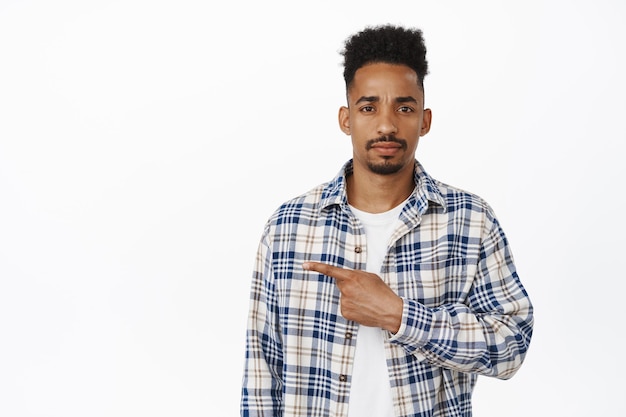 Sad african american guy, young man pointing finger left, frowning and looking let down, upset and disappointed about smth, showing bad promo text, white background.
