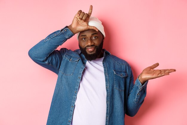 Sad african-american man showing loser sign on forehead and staring at camera, standing over pink background