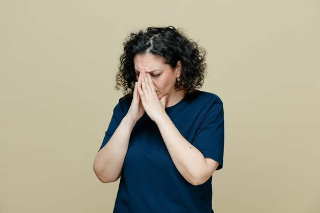 грустная и взволнованная женщина средних лет в футболке держит руки вместе на носу, глядя вниз, изолированная на оливково-зеленом фоне