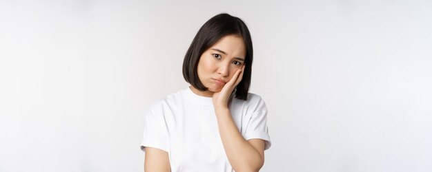 Sad asian girl looking upset and lonely sulking and frowning standing against white background in casual tshirt