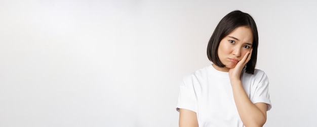 Sad asian girl looking upset and lonely sulking and frowning standing against white background in casual tshirt