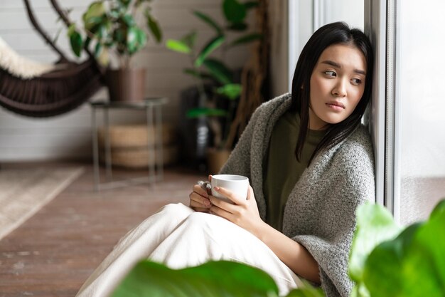Sad asian woman looking outside window sitting on floor with miserable upset face drinking coffee at...