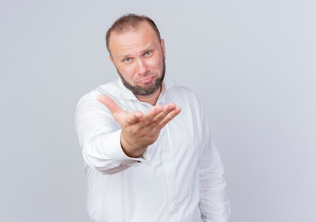 Triste uomo barbuto che indossa una camicia bianca confuso con il braccio fuori come chiedere in piedi sul muro bianco