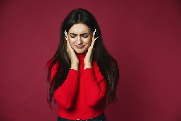 Sad beautiful brunette caucasian girl dressed in red pullover with annoying headache is holding head in hands