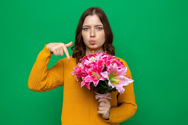 Sad beautiful young girl on happy woman's day holding and points at bouquet isolated on green wall