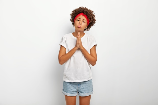 Бесплатное фото Грустная женщина-попрошайка с афро-волосами держит руки в молитвенном жесте, поджимает губы и просит огромную услугу, помогая в беде, носит белую удобную футболку и джинсовые шорты, требует извинений