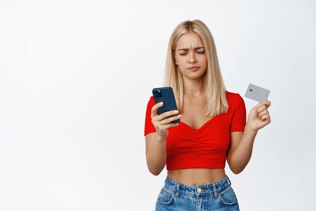 슬픈 금발 10대 소녀는 흰색 배경 복사 공간 위에 서 있는 온라인 구매에 문제가 있는 휴대폰과 신용 카드를 보유하고 있습니다.