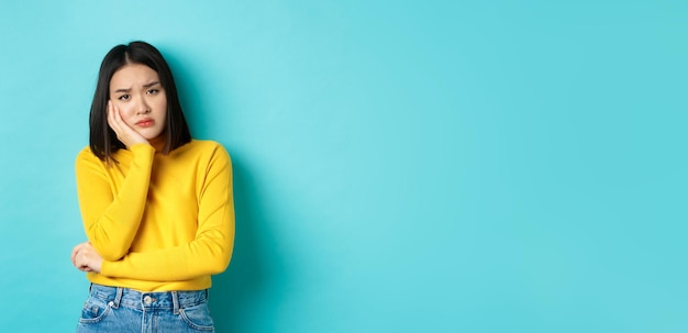 Free photo sad and bored asian girl looking reluctant and unamused at camera leaning face on hand standing over