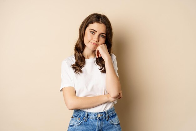 Sad and bored brunette girl looking unamused, uninterested, standing over beige background