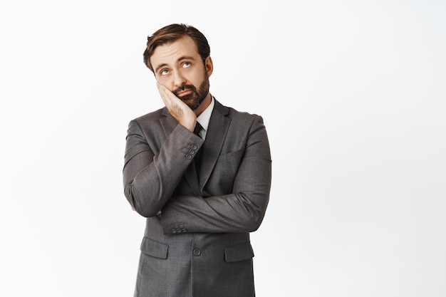 Sad and bored businessman in suit looking up roll eyes and lean on hand face palm standing unamused and reluctant over white background