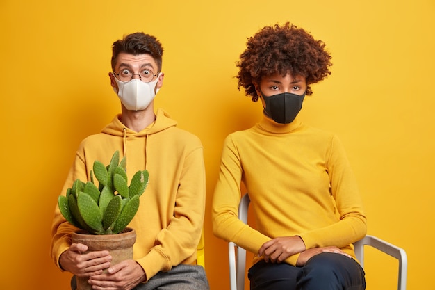 sad bored woman and shocked guy sit together on chairs wear medical masks carry potted cactus