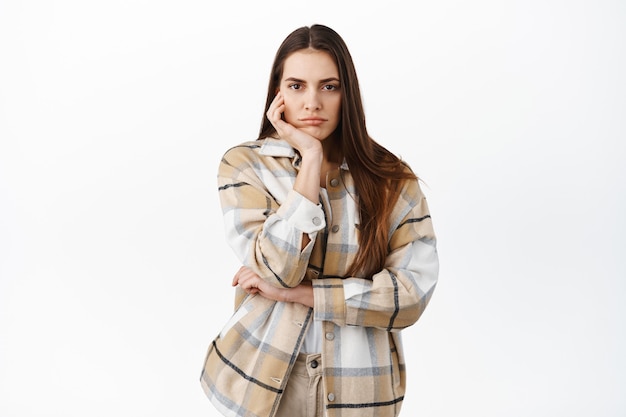 Sad or bored woman stare indifferent at front, no emotions poker face, lean head on palm and looking at something boring, standing against white wall