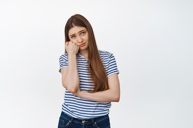 Sad and bored young woman looking up, leaning head on arm, standing against white background unamused