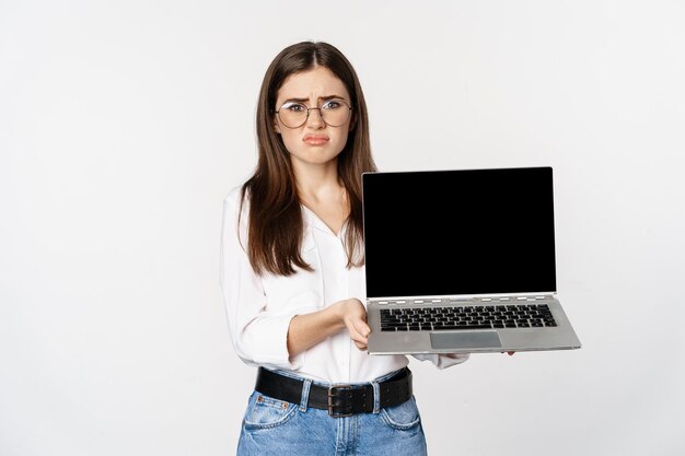 Sad brunette girl, student showing laptop screen and grimacing upset, standing disappointed against white background