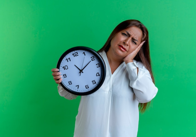 Sad casual caucasian middle-aged woman holding wall clock and putting hand on cheek isolated on green wall