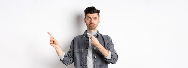 Free photo sad caucasian guy pointing fingers right and pouting looking hesitant standing on white background
