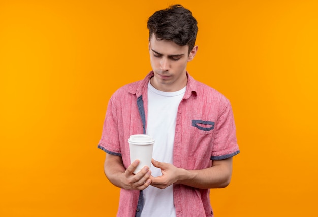 Sad caucasian young guy wearing pink shirt holding cup of coffee on isolated orange background