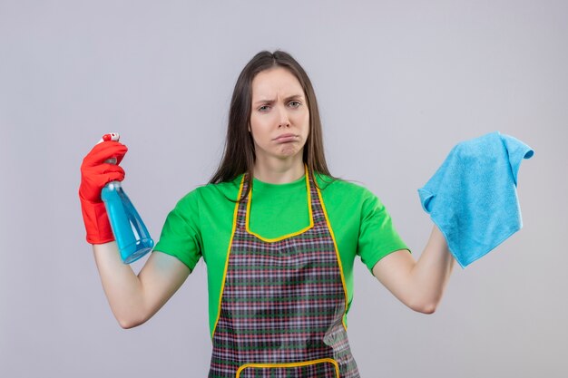Sad cleaning young girl wearing uniform in red gloves holding cleaning spray and rag on isolated white background