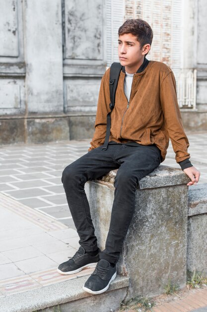 Sad contemplated teenage boy with shoulder bag sitting on wall