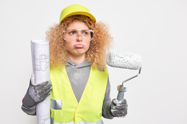 悲しい縮れ毛の女性建築家は、プロの住宅建設労働者がローラーを保持し、青写真がプロジェクトのいくつかの間違いを認識している保護ヘルメットの作業服を着ています。産業の構築