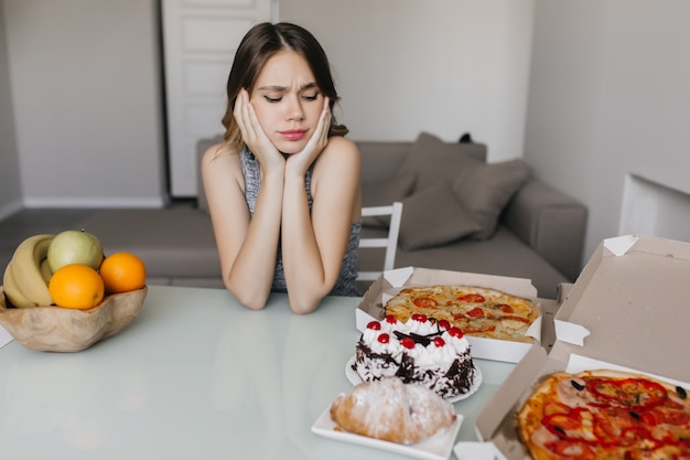 Sad curly woman looking at cake during diet. blonde gorgeous female model posing with fruits and pizza.