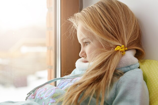 Sad cute female child with blonde hair sitting on windowsill