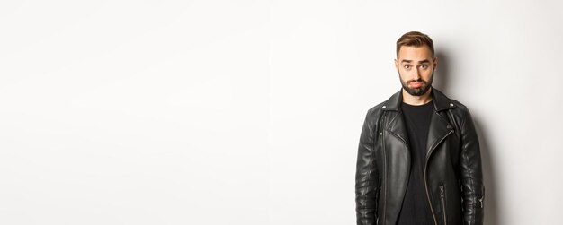Sad and cute guy in leather black jacket pouting looking silly at camera standing against white back