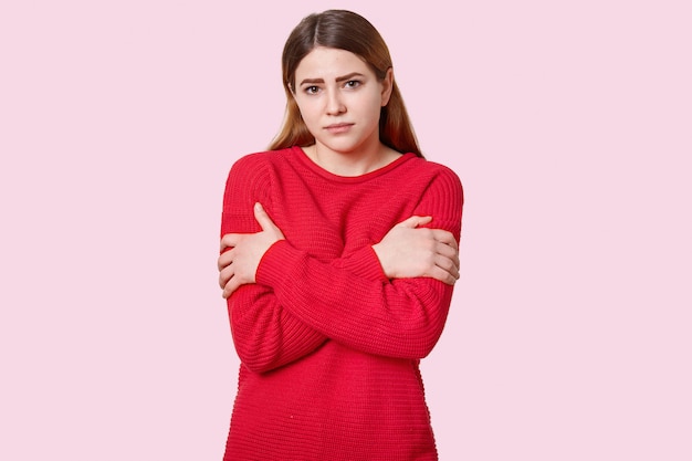 Sad dark haired woman crosses hands over chest, has make up, dressed in loose red sweater, models over pink