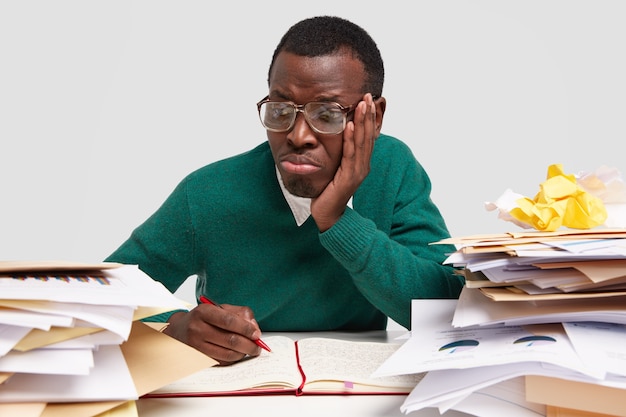 Sad dark skinned male hipster feels overworked, overloaded, notes ideas in notebook, purses lips in displeasure, wears big square glasses for good vision