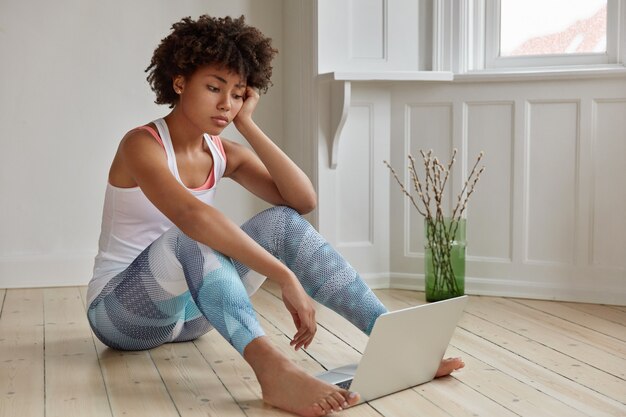 Sad dark skinned student dressed in casual outfit, sits bare foot on wooden floor in empty room with laptop compute