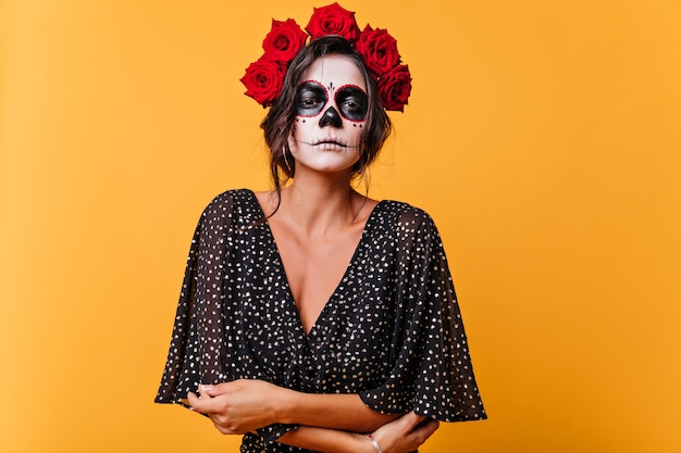 Free photo sad dead bride with zombie makeup standing on yellow wall. elegant latin woman with roses in hair preparing for halloween.