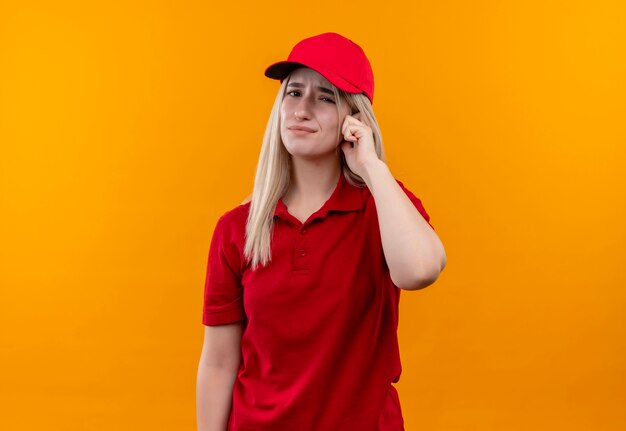 Грустная молодая девушка из службы доставки в красной футболке и кепке положила палец на голову на изолированном оранжевом фоне