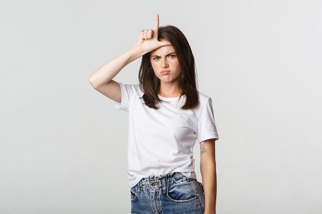 Sad and disappointed brunette girl showing loser gesture and sulking, white.