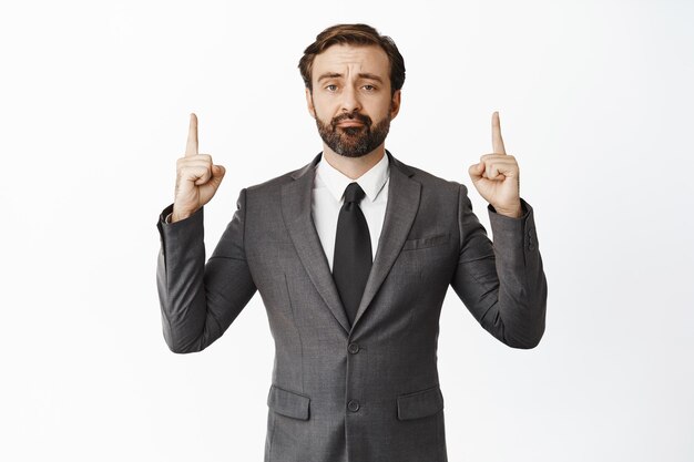 Sad and disappointed businessman in suit pointing fingers up pulling upset face and grimacing showing upsetting news white background