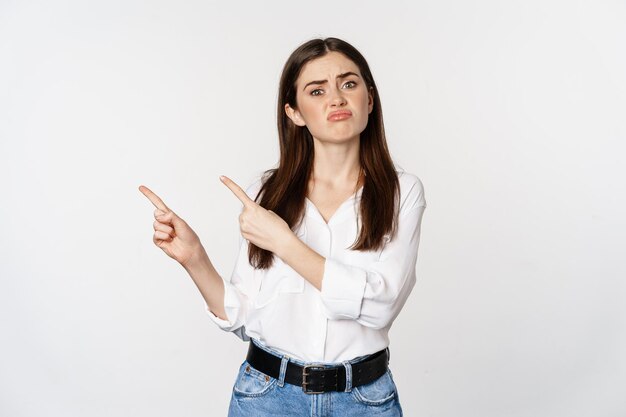 Sad and disappointed cute woman, gloomy girl pointing fingers at logo, complaining, sulking upset, standing in blouse over white background