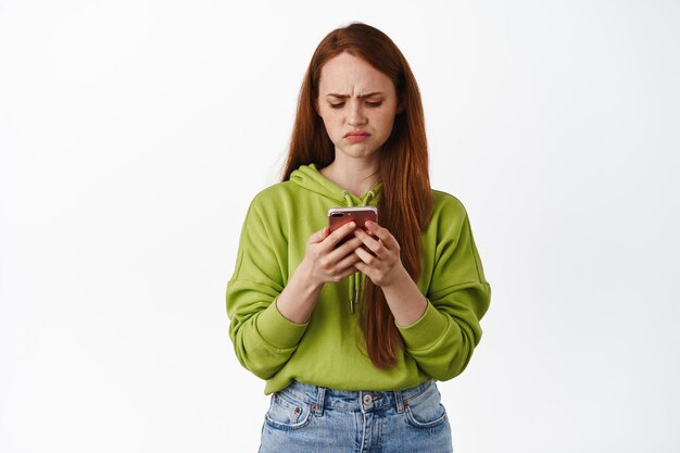 Sad and disappointed ginger teen girl looking upset at her phone, staring at smartphone with regret or jealousy, standing against white background. Technology concept