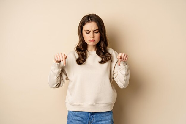 Sad and disappointed girl pointing looking down with upset grimace moody face standing over beige ba...