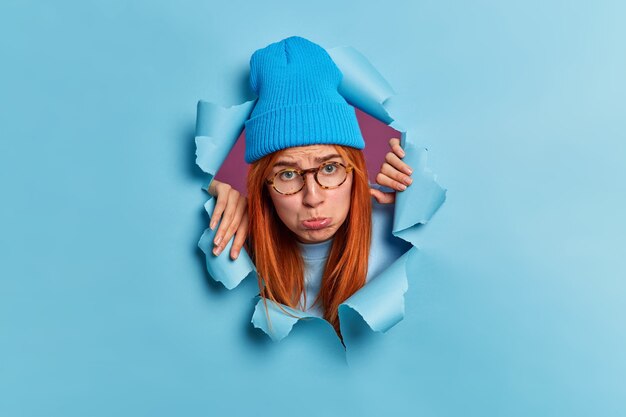 Sad disappointed redhead teenage girl purses lips and looks with sullen face expression wears blue hat and spectacles looks through hole of torn paper