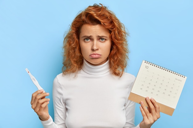 Free photo sad disappointed redhead woman holds pregnancy test and menstruation calendar