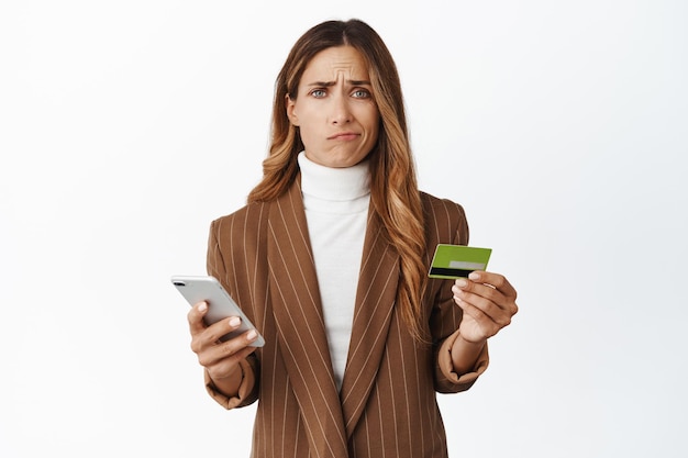 Sad and disappointed woman holding credit card and smartphone bad user experience with mobile app problem with card register online white background