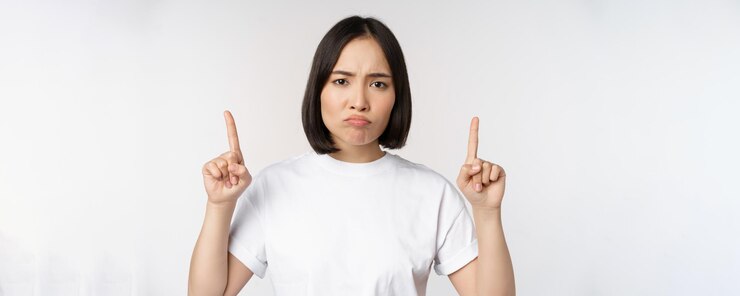 無料Photo 悲しくて失望した若いアジアの女性が指を上に向けて動揺した表情の白いスタジオの背景で広告を表示