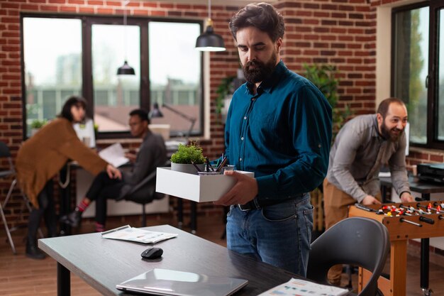 Sad dismissed man holding box with belongings after being fired while his collegues looking at hom working in startup office. Businesspeople brainstorming ideas developing marketing project