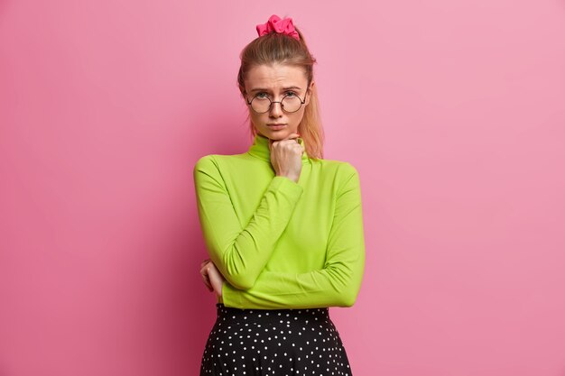 Sad displeased European woman with pony tail keeps hand under chin and looks unhappily