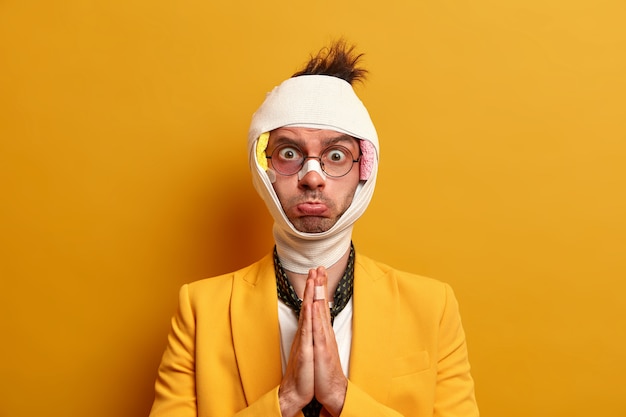 Free photo sad displeased man keeps palms pressed togehter and asks for help, has bandage over head, broken nose, bruises under eyes, swollen face poses against yellow wall. victim of accident or injury