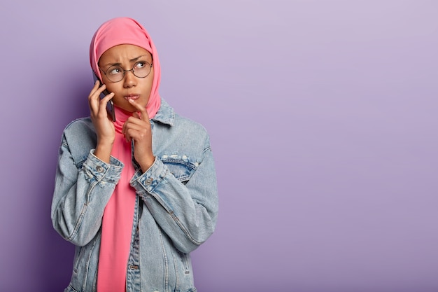 Sad dissatisfied dark skinned woman wrapped in pink hijab, wears denim jacket, round spectacles