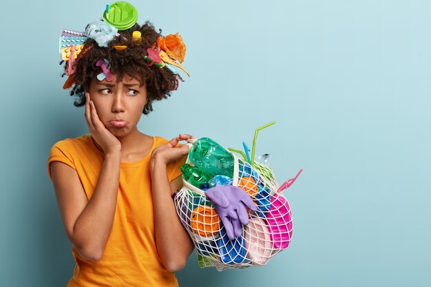 悲しい不満の女性は下唇を財布に入れ、怒って脇に見え、プラスチック廃棄物の入ったネットバッグを持ち、領土をきれいにし、環境にやさしく、環境を汚染する人々に腹を立て、オレンジ色のTシャツを着ています