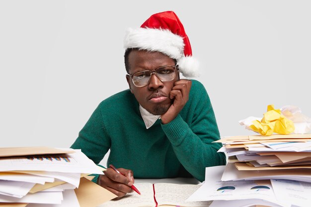 Sad dissatisfied man purses lips, keeps hand on cheek, wears Santa Claus hat, works hard before celebrating winter holidays