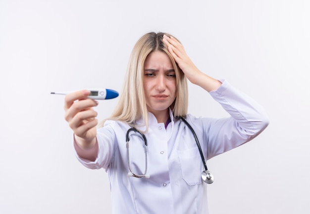 Stetoscopio da portare della giovane ragazza bionda di medico triste e termometro della tenuta dell'abito medico alla macchina fotografica mise la sua mano una testa su fondo bianco isolato